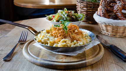 Traditionelle Käsespätzle mit Röstzwiebeln, serviert mit gemischtem Salat und Brezeln auf einem rustikalen Holztisch.