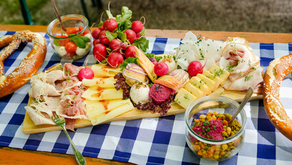 Traditionelles Brotzeitbrett mit Käse, Schinken, Obatzda, Radieschen und Brezeln auf blau-weißer Tischdecke im Biergarten.