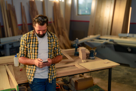 A carpenter in a workshop looking at his smartphone while leaning against a workbench.