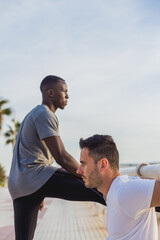 Two men, one Caucasian and one Black, stretching together before a workout on the beach at sunset