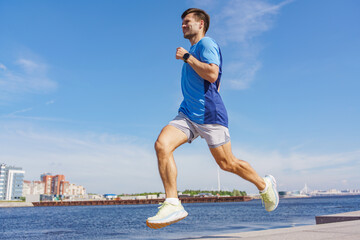 Young man jogging along the riverbank on a sunny day near the city skyline in summer