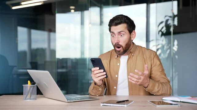 Amazed happy businessman looks at mobile phone screen sitting at workplace in office. Excited shocked man read great news, message or saw pleasant content. Male celebrates success looking at camera