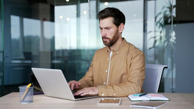 Frustrated businessman having difficulty making transaction using laptop while sitting at desk at workplace in a modern business office. Confused male worker having problems working online on computer