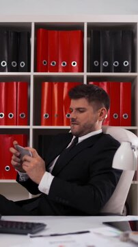 Ultra HD Young attractive businessman playing in game on the mobile iphone in modern cool office on armchair with lamp computer digital pad, keyboard. teal and orange. Vertical Shot.