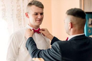 Two men are getting ready for a wedding. One man is helping the other tie his red bow tie