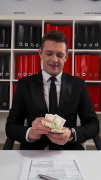 4k uhd POV Young attractive business man in black suit and tie counting dollars with smile. The promise that the work and servise will be done. Hide money in pocket. Office indoor. Vertical Shot.