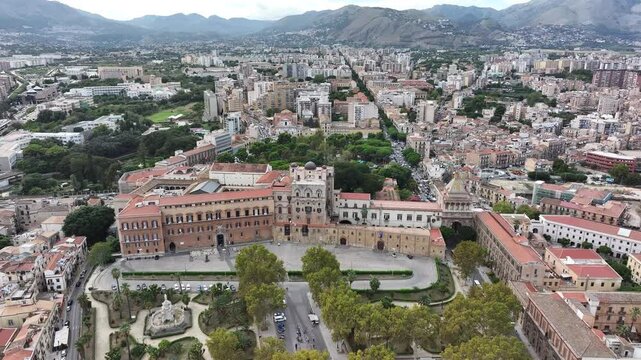 An aerial view of Palermo, Sicily, showcasing the historic Palazzo dei Normanni with its distinctive architecture, surrounded by lush greenery, city streets, and distant mountains under a cloudy sky.
