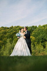 A bride and groom are standing in a field, with the bride holding a bouquet and the groom holding her hand