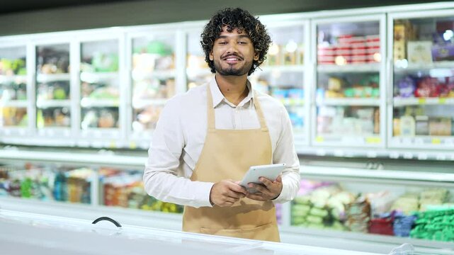 Portrait young staff man salesman in apron looking at camera in grocery store supermarket. Handsome male worker seller or small business owner indoor. food groceries market smile using a tablet