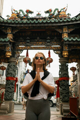 Young Blonde Woman in Sunglasses Practicing Prayer Pose in Front of Traditional Asian Temple Gate.