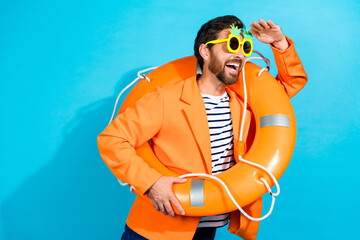 Charismatic young man wearing orange jacket and novelty sunglasses holding a lifebuoy, joyfully...