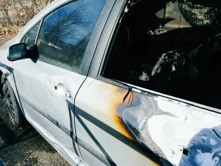 Close-up of a silver car with visible fire damage, including charred and burnt areas on the door and window, parked outdoors on a sunny day.