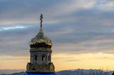 A golden Orthodox church dome against a dramatic sunset sky with mountains in the background.