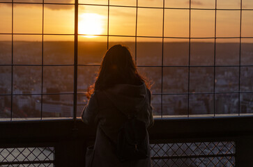 A silhouette of a woman gazing at the sunset from a high vantage point, framed by a wire fence. The golden hour light emphasizes her hair and the tranquil urban landscape 