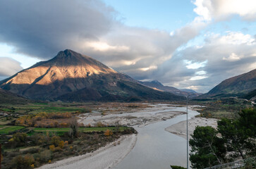 A view of the Vjosa valley in Tepelenë Albania with the Vjosa river winding through it, framed by majestic mountains. The sky is filled with dramatic clouds, creating a stunning natural scene.