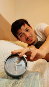 Vertical video of a young man lazily turning off his alarm clock in the morning and snuggling back into bed, concept of morning laziness, cozy bedroom routine, sleep habits, and humorous daily life
