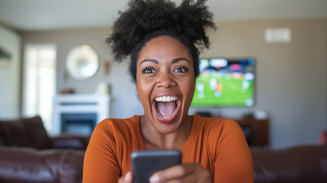 Enthusiastic african american woman cheering during soccer match, holding smartphone and celebrating victory while watching game at home