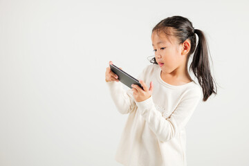 Fototapeta premium Happy Asian portrait cute young kid girl smiling excited using smart mobile phone studio isolated on white background, Thai kindergarten child surprised holding reading message gesture on smartphone