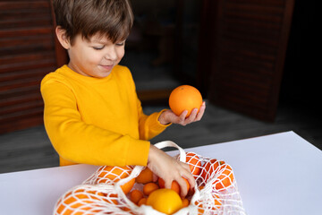 A child has picked an orange and reaches for a tangerine from a mesh bag.