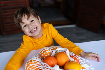 Fototapeta premium A child, standing in front of the table, smiles as they hug a bag filled with citrus fruits.