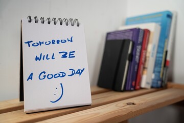 tekst motywacyjny" Tomorrow will be a good day zapisany w notesie stojącym półce z książkami © siwyk