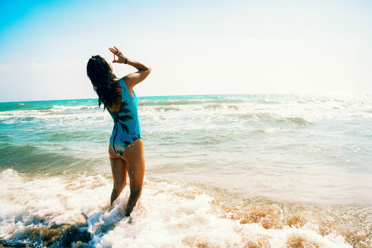 Girl in blue swimsuit on sea coast adjusts hair and enjoys rest. View from back. Generated image