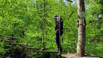 Guy Performs a Complex Obstacle Course Attraction Suspended in the Treetops. An Aerial Adventure Course is an Obstacle Course Set High up in the Trees. Courses Get More Difficult and Higher.