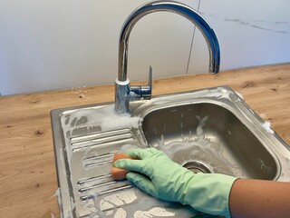 A hand in a rubber glove washes the kitchen sink with a sponge, covered in foam from detergent....