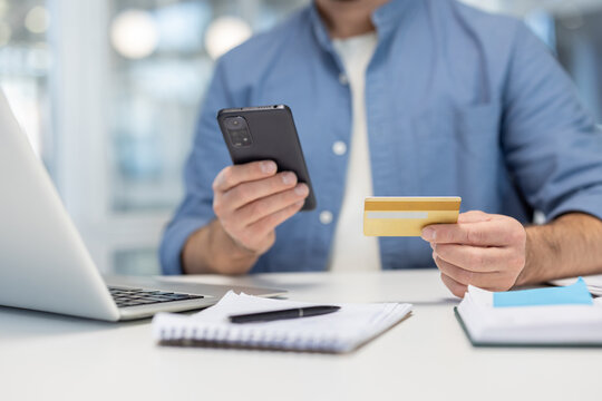 Man hands holding phone and bank credit card close up, businessman in casual clothes working inside office with laptop and documents. - Powered by Adobe