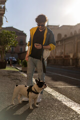 Stylish man checking phone while walking French Bulldog at sunset