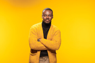 Confident man in a yellow cardigan posing with folded arms against a warm golden background in a studio setting