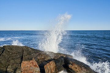 A wave crashing into a large rock on the Rhode Island seashore. It is a beautiful clear day, and the ocean is a beautiful deep blue.