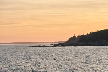The sky is a beautiful yellow at sunset here in Acadia National Park. The rocky coastline is a silhouetted against the sky, and the ocean is calm. This image includes copy space for text.