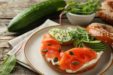 Delicious bagel with salmon, cream cheese, cucumber, avocado and pumpkin seeds on wooden table, closeup