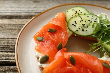 Delicious bagel with salmon, cream cheese, cucumber, avocado and pumpkin seeds on wooden table, closeup