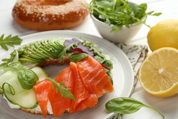 Delicious bagel with salmon, cream cheese, cucumber and avocado on white wooden table, closeup