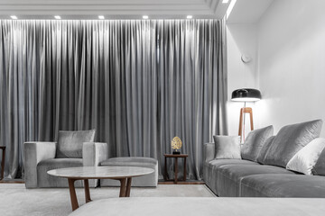 stylish large living room in white and gray tones, gray sofa, armchair, round table, wooden coffee table, carpet, floor lamp, gray curtains on windows. Illuminated by lamp. Space for relaxation
