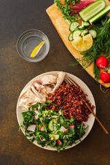 Delicious healthy breakfasts, chicken breast with red rice and salad mix with fresh seasonal organic vegetables and microgreens, top view of a plate with food and a cutting board with vegetables