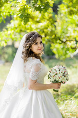 A woman in a white dress is holding a bouquet of flowers. She is wearing a veil and has her hair in a bun. The scene is set in a park, with trees in the background. The woman is happy and content
