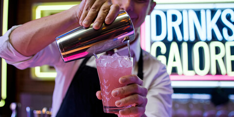 Bartender Pouring a Refreshing Pink Cocktail. A smiling bartender expertly pours a vibrant pink cocktail over ice at a stylish bar, with a neon sign glowing in the background.