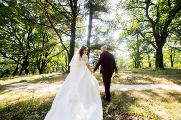 A bride and groom are walking through a forest, hand in hand. The bride is wearing a white dress and the groom is wearing a red suit. The scene is peaceful and romantic
