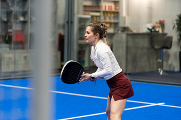 Woman playing padel in a blue grass padel court indoor - Young sporty woman padel player hitting ball with a racket