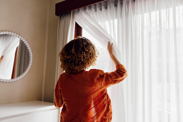 A woman with curly hair wearing an orange blouse opens sheer curtains, letting sunlight in, in a...