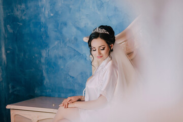 A woman in a white dress sits on a table with her hands on her lap. She is wearing a tiara and has...