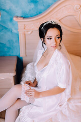 A woman in a white gown sits on a bed with her legs crossed. She is wearing a white veil and a tiara