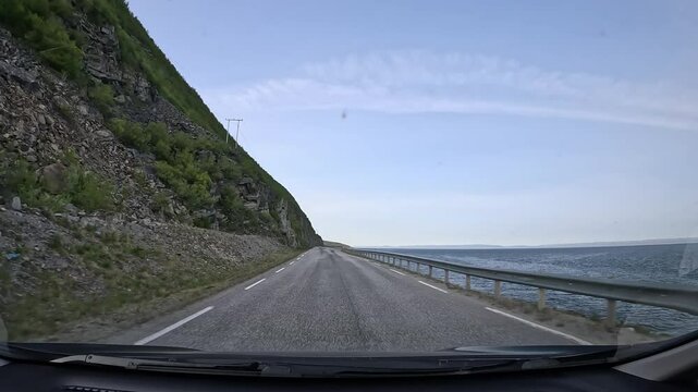 Driving along Norway's northern tundra on E69 highway. The sea stretches on the right, while a steep hill and rocky cliffs with low shrubs rise on the left. A scenic and tranquil coastal drive.