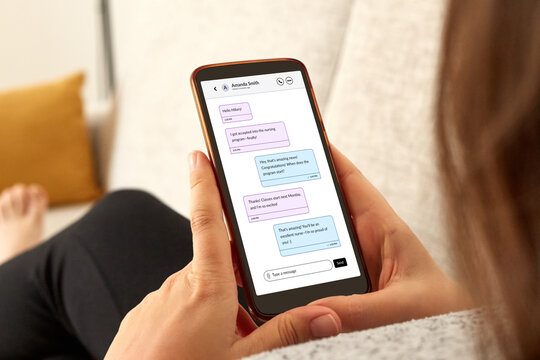 Close-up photo of a girl seen from behind, sitting on her home sofa and using an instant messaging app on her smartphone, symbolizing online chatting.