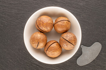 Macadamia nuts in shell with ceramic saucer on slate stone, top view.