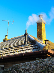 Naklejka premium A newly fitted flue pipe from a burning heating boiler, expelling fumes through a frosty domestic house roof, next to a chimney stack and TV aerial, on a cold winter morning, against a blue sky.