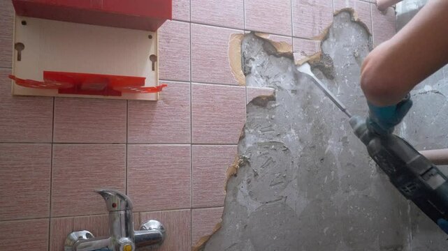 Construction worker is removing tiles from bathroom wall using jackhammer. Wall is partially demolished, revealing concrete underneath
